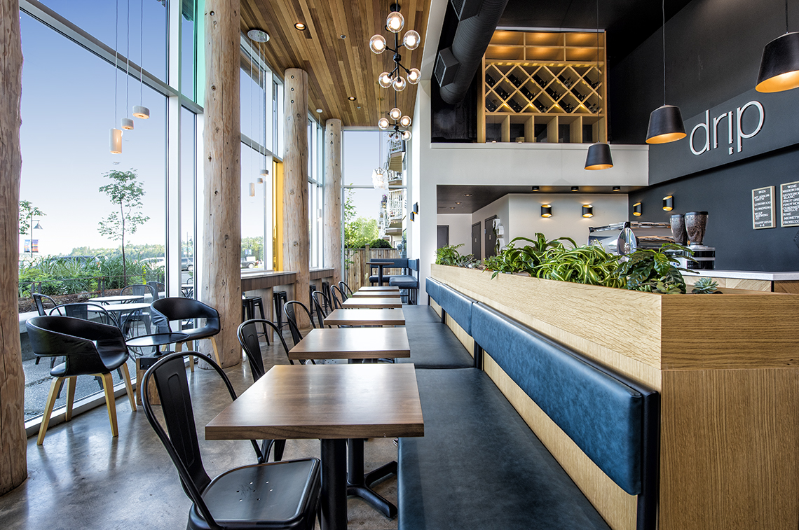 Interior of Drip Coffee | Social. Modern café interior with tall wooden columns, large windows, and stylish pendant lights. The seating area features wooden tables, blue bench seating, and black metal chairs. The coffee counter is lined with greenery, and 'drip' is written on the wall in bold, minimalist font.