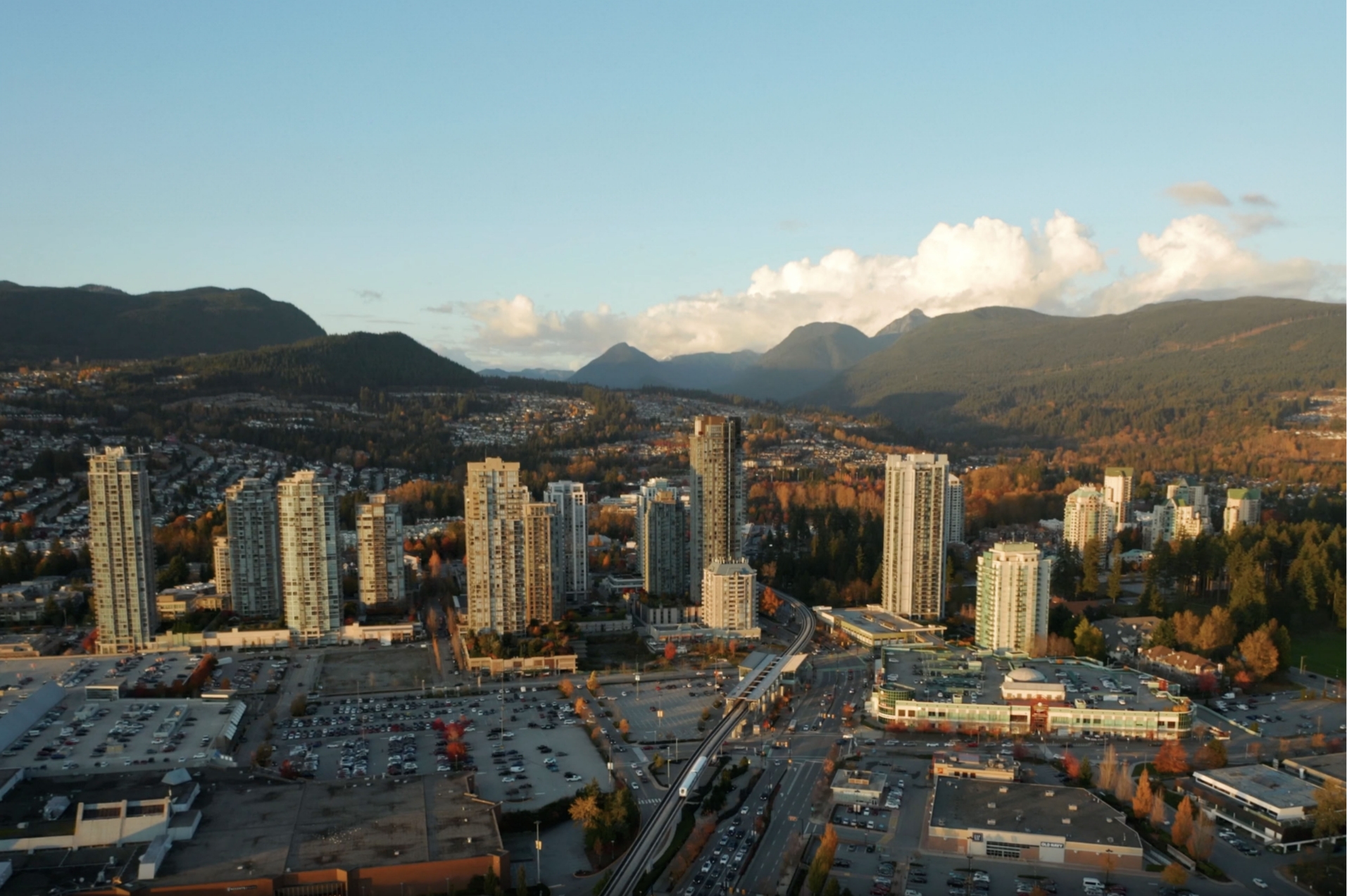 A scenic aerial view of Coquitlam's skyline with high-rise buildings in the foreground and a backdrop of forested hills and mountains. The cityscape includes parking lots, roads, and surrounding neighborhoods, with a clear, bright sky overhead.